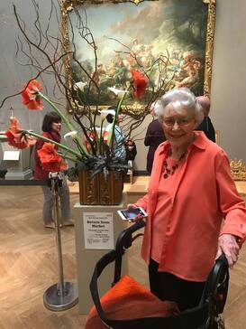 Elizabeth Sherrill wearing a beautiful deep orange shirt standing in front of a flower arrangement at a museum. She is smiling. 